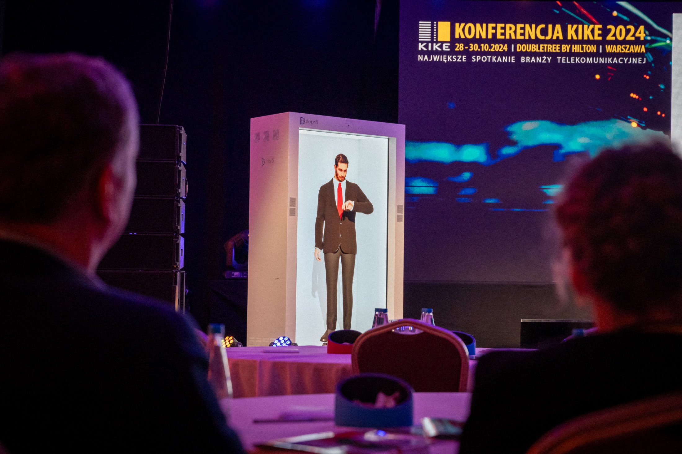 The avatar at life size in a holographic display cabinet on the conference stage, idling between conversations